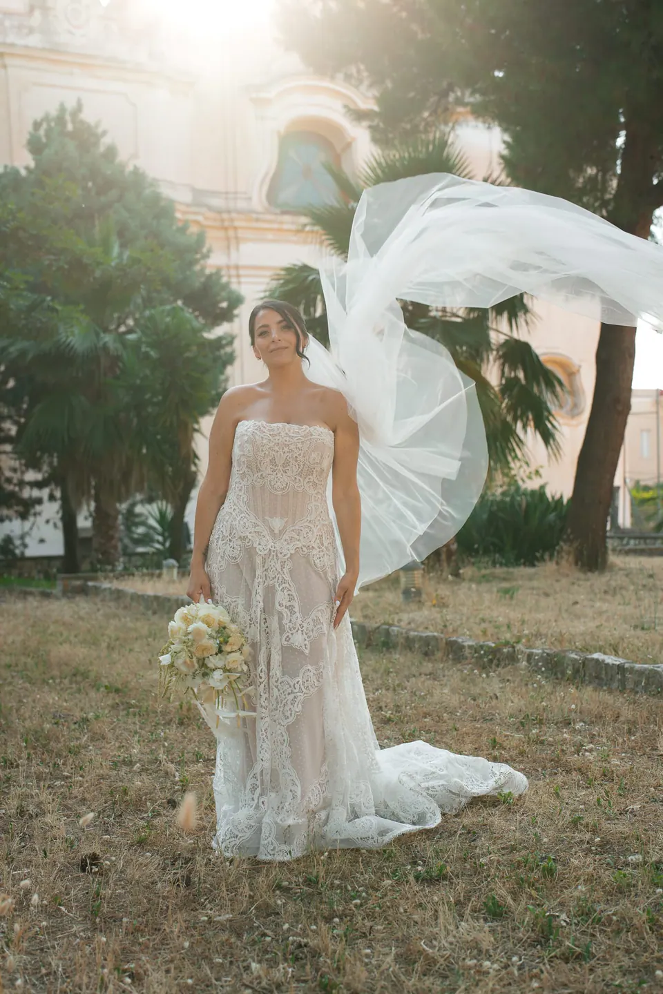 Bride on the lawn with her bouquet, the veil lifted high by the wind.