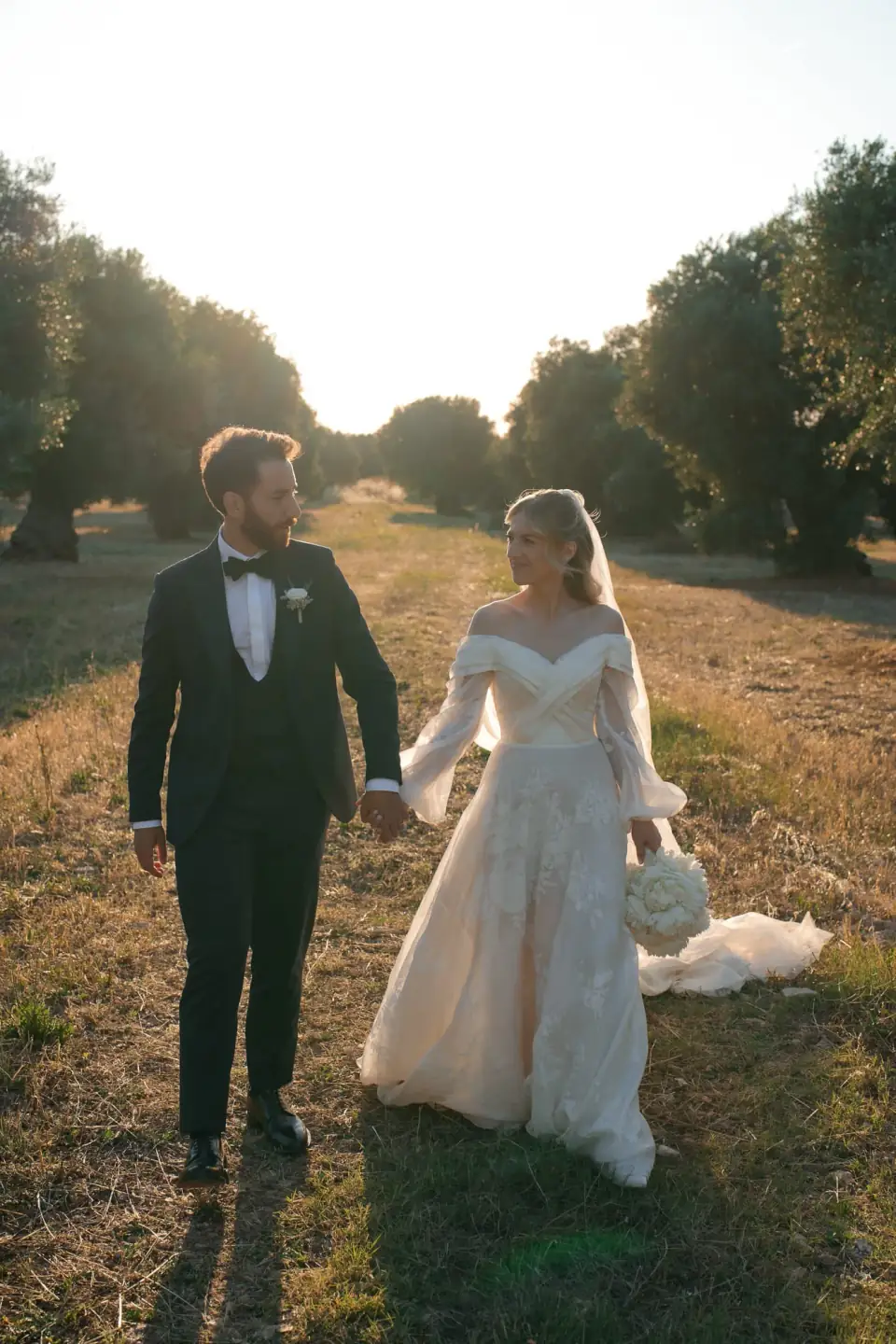 Bride and groom walking hand in hand through the olive grove of Masseria Montalbano at sunset.