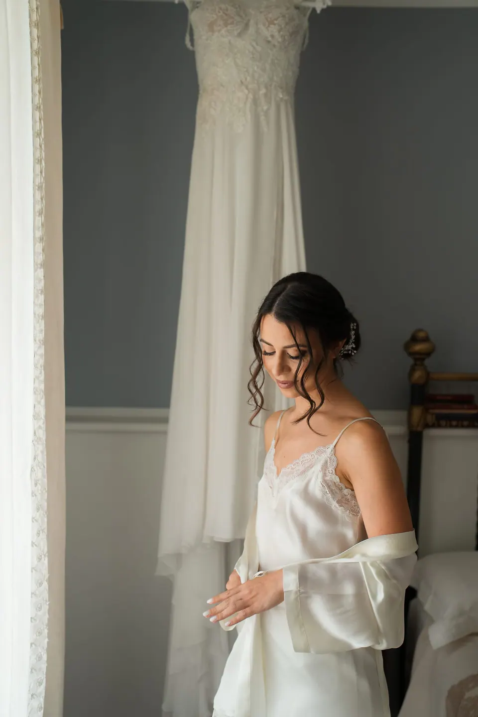 Bride in a silk slip by the window, the wedding gown hanging beside her.