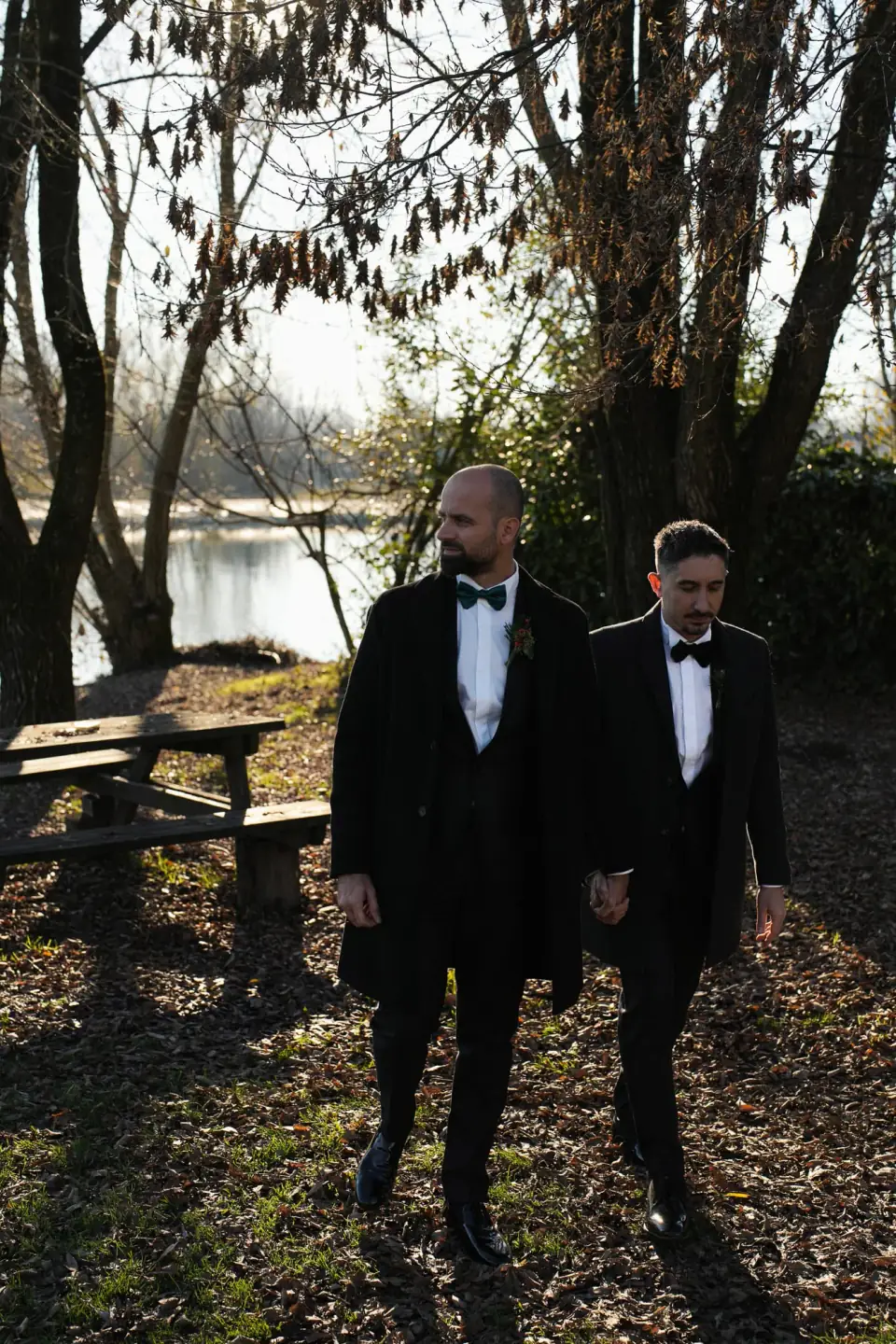 The two grooms walking side by side under the trees by the river at Castello Visconteo.