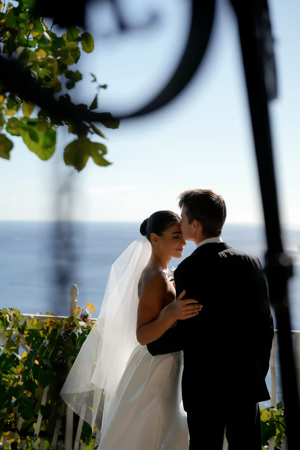 Bride and groom embracing beneath a wrought-iron lamp, the sea beyond.