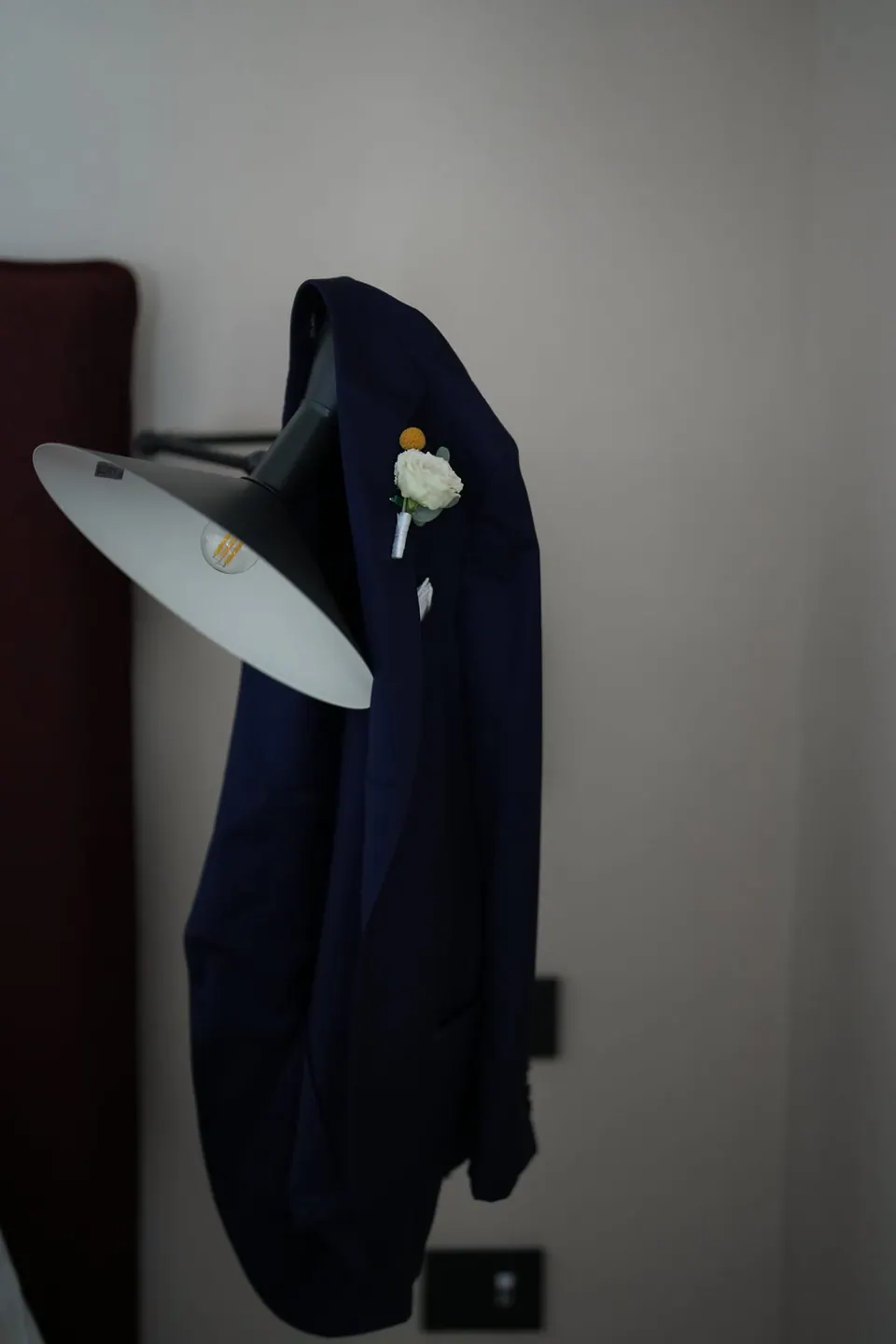 Groom's navy jacket with boutonniere hanging beside a lamp.