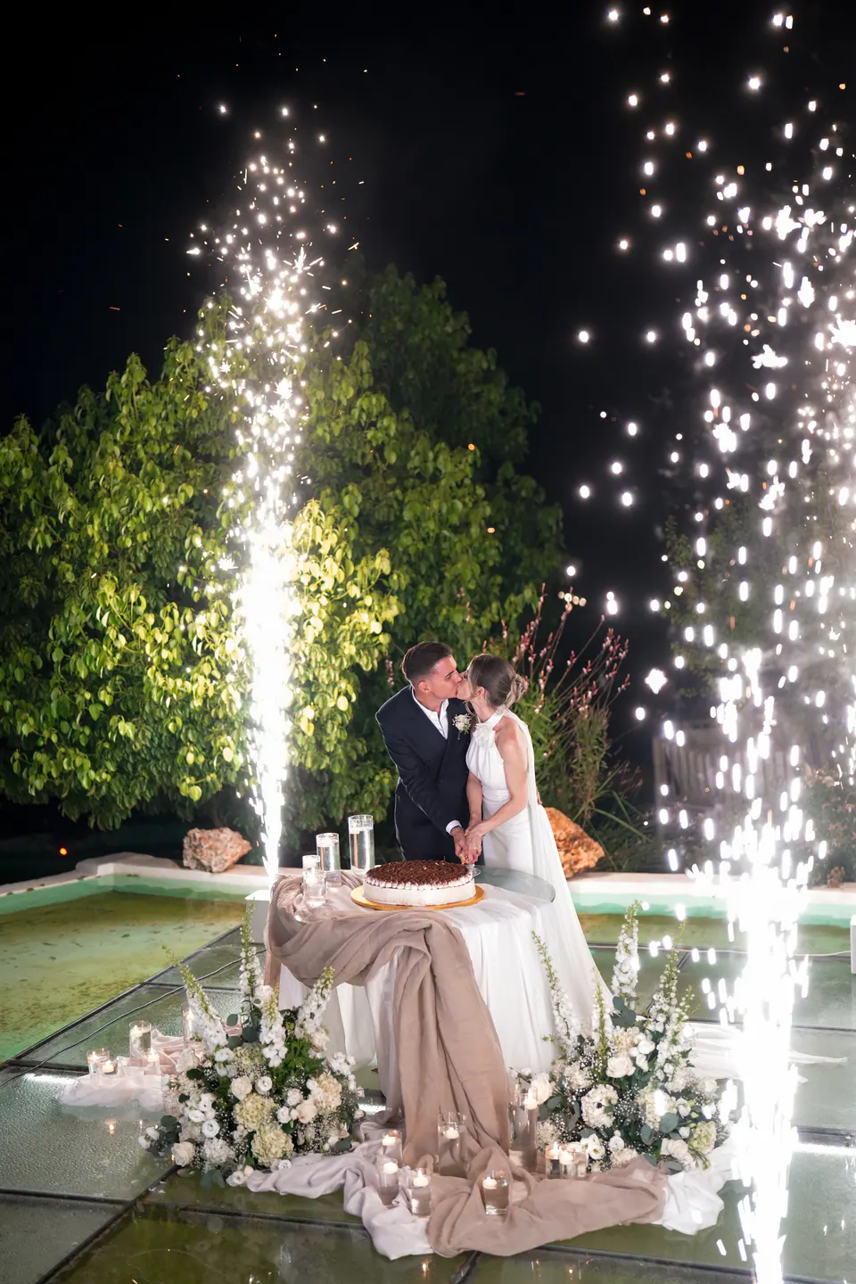 Bride and groom kissing by the cake as fountains of sparks rise into the night sky.