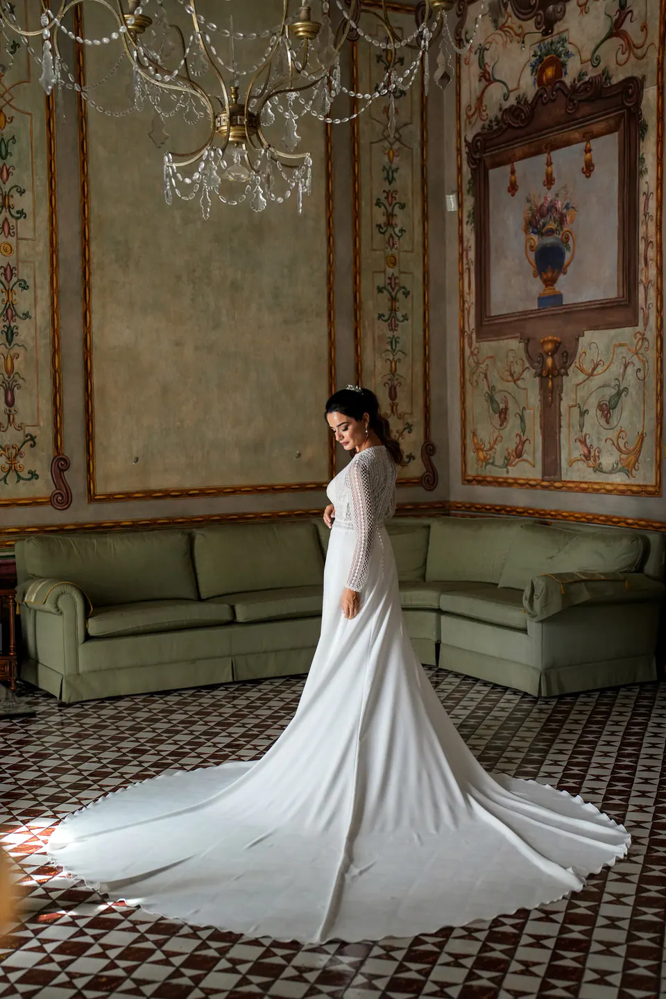 Bride in a frescoed palace room beneath a crystal chandelier, her long train spread on the floor.